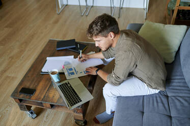 Male entrepreneur writing on paper while sitting with laptop at home - KIJF03396