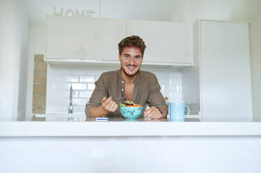 Smiling man with salad bowl leaning over kitchen counter at home - KIJF03399