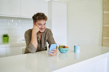 Smiling man using smart phone while leaning on kitchen counter at home - KIJF03401