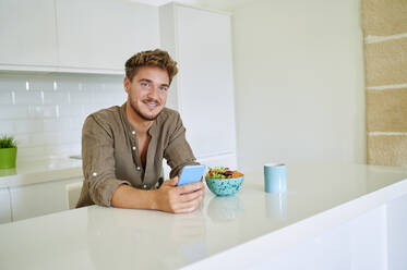 Young man with smart phone standing by counter in kitchen at home - KIJF03402