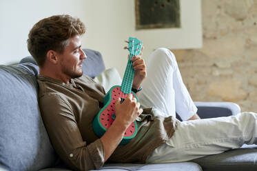 Young man playing ukulele on sofa in living room at home - KIJF03413