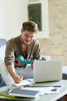 Junger Mann schreibt auf Papier, während er im Wohnzimmer auf einem Laptop online Ukulele lernt - KIJF03416