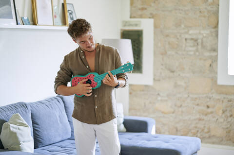 Male musician playing ukulele while standing in living room at home stock photo