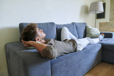 Young man relaxing on sofa with hands behind head in living room - KIJF03421