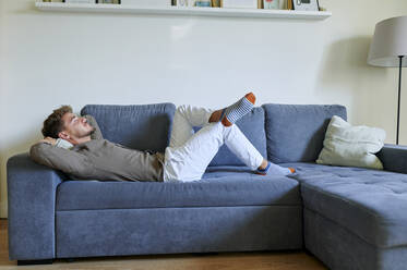 Man resting on sofa with hands behind head in living room at home - KIJF03422