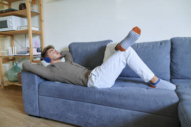 Young man with headphones listening music while lying on sofa at home - KIJF03423