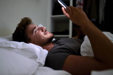 Caucasian man using smart phone while lying on bed in bedroom at night - KIJF03434