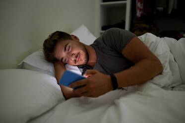 Junger Mann, der nachts auf dem Bett liegend eine SMS über sein Mobiltelefon schreibt - KIJF03435
