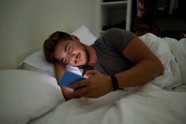 Young man text messaging through mobile phone while lying on bed at night - KIJF03435