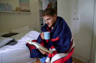 Junger Mann, eingewickelt in eine Decke, liest ein Buch mit einem Kaffeebecher im Schlafzimmer zu Hause - KIJF03436