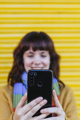 Young woman taking selfie on smart phone against yellow shutter - MGRF00070