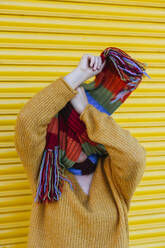 Junge Frau bedeckt ihr Gesicht mit einem Schal vor einem gelben Fensterladen - MGRF00072