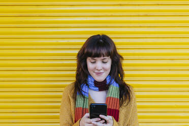 Smiling woman text messaging on smart phone while standing against shutter - MGRF00074