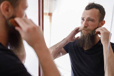 Mittlerer erwachsener Mann, der seinen Bart mit einem Kamm bürstet, während er einen Spiegel im Wohnzimmer betrachtet - IFRF00121