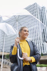 Lächelnde Geschäftsfrau mit Regenschirm, die in der Stadt während der Regenzeit wegschaut - IFRF00126