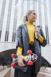 Blonde Frau mit Skateboard, die wegschaut und einen Regenschirm in der Hand hält - IFRF00128