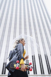 Berufstätige Frau mit Skateboard und Regenschirm auf dem Weg zur Arbeit in der Regenzeit - IFRF00130