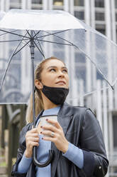 Nachdenkliche Geschäftsfrau mit Gesichtsmaske hält Regenschirm und Einwegbecher in der Stadt während der Regenzeit - IFRF00131
