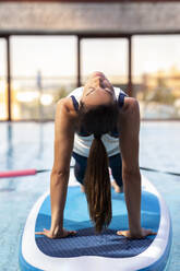Weiblicher Yogalehrer in Brückenposition auf dem Paddleboard über dem Schwimmbad - JCCMF00086