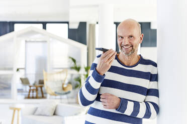 Man smiling while talking on mobile phone standing at home - FMKF06757