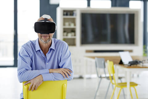 Lächelnder Geschäftsmann, der ein Virtual-Reality-Headset benutzt, während er sich zu Hause an einen Stuhl lehnt, lizenzfreies Stockfoto
