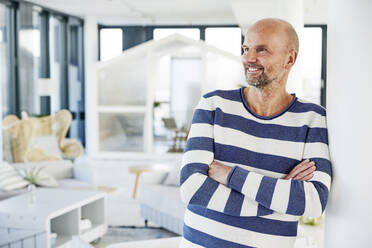 Smiling man with arms crossed looking away while standing at home - FMKF06763