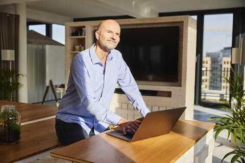 Smiling businessman looking away while using laptop sitting at home stock photo