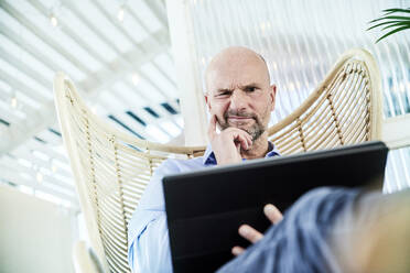 Verwirrter Geschäftsmann bei der Arbeit an einem digitalen Tablet, während er zu Hause auf einem Stuhl sitzt - FMKF06782