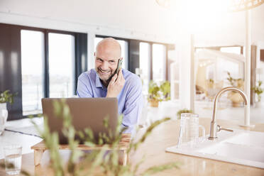 Unternehmer mit Laptop, der im Stehen zu Hause mit seinem Handy telefoniert - FMKF06792