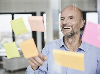 Smiling businessman reading adhesive note on glass wall - FMKF06801