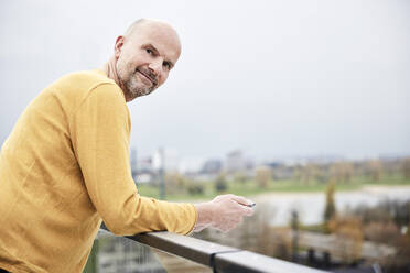 Smiling man using mobile phone while standing on rooftop - FMKF06805