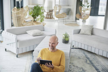 Älterer Mann, der ein digitales Tablet benutzt, während er zu Hause auf dem Boden sitzt - FMKF06812