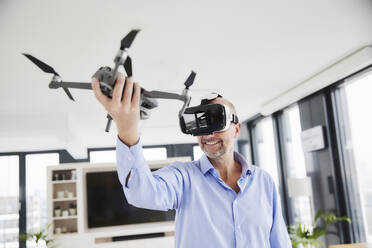 Entrepreneur wearing virtual reality headset holding quadcopter while standing at home - FMKF06822