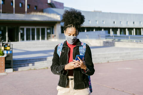 Student wearing protective face mask using smart phone while standing at city street stock photo