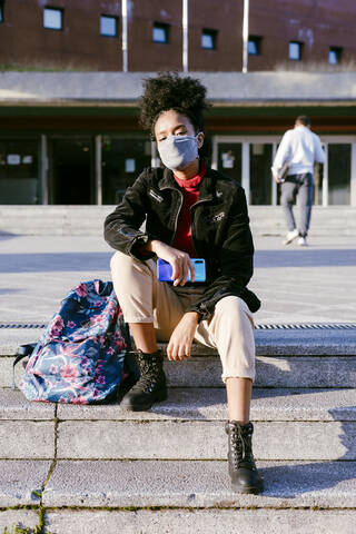 Junge Frau mit Gesichtsschutzmaske auf einer Treppe in der Stadt sitzend, lizenzfreies Stockfoto
