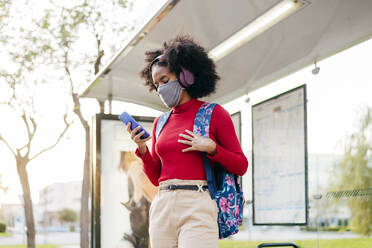 Young woman wearing headphones and protective face mask using smart phone while standing at bus stop - MARF00005