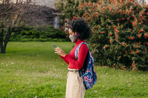 Student wearing headphones and protective face mask using smart phone while standing outdoors stock photo