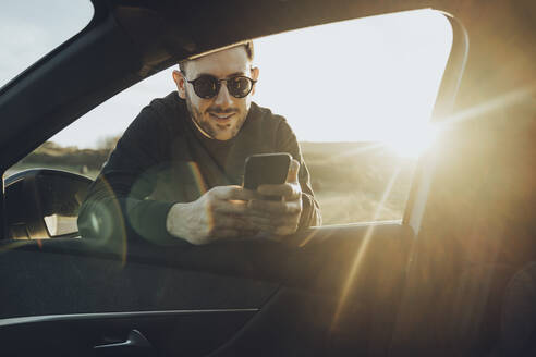 Gut aussehender junger Mann, der sein Smartphone benutzt und sich bei Sonnenuntergang an ein Autofenster lehnt - MTBF00729