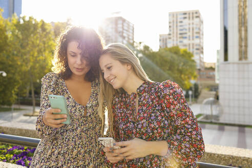 Freundinnen benutzen ein Smartphone an einer Stützmauer in der Stadt - IFRF00137