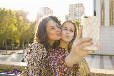 Female friends puckering while taking selfie through smart phone on sunny day - IFRF00139