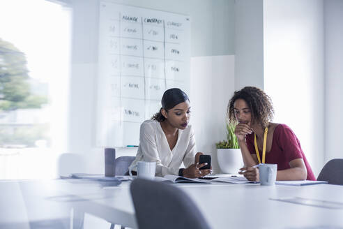 Female colleagues using smart phone while sitting at desk in office - AJOF00792