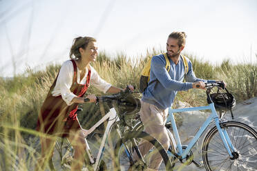 Smiling girlfriend and boyfriend walking with bicycles at beach - UUF22191