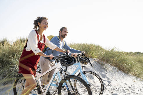 Lächelndes Paar schaut weg, während es mit Fahrrädern am Strand spazieren geht - UUF22192