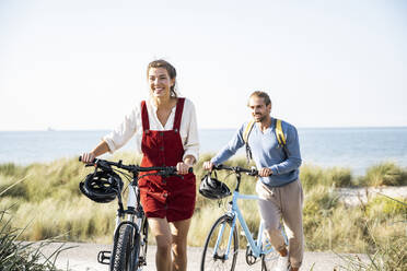 Smiling man and woman walking with bicycles at beach - UUF22199