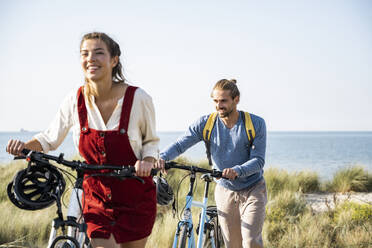 Smiling young man and woman walking with bicycles against sea - UUF22200