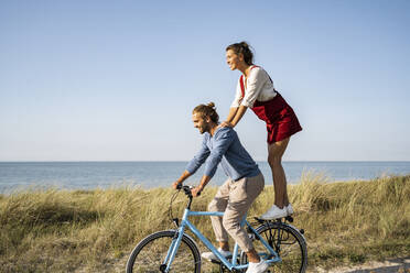 Smiling woman enjoying ride with man while standing on bicycle against clear sky - UUF22210