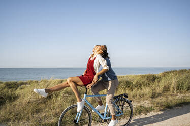 Happy girlfriend sitting on handle while enjoying bicycle ride with boyfriend against clear sky - UUF22212