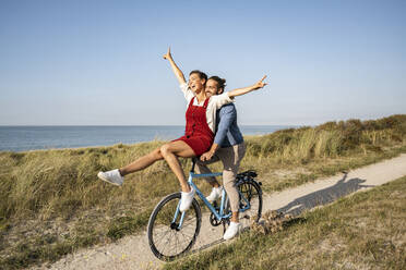 Woman with hand raised sitting on handle while enjoying bicycle ride with boyfriend against clear sky - UUF22213