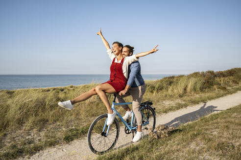 Frau mit erhobener Hand auf dem Lenker sitzend, während sie eine Fahrradtour mit ihrem Freund gegen den klaren Himmel genießt - UUF22213