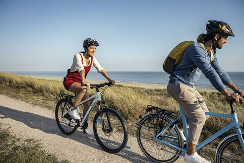 Junge Frau lächelt beim Fahrradfahren mit einem Mann auf dem Fußweg gegen das Meer - UUF22223